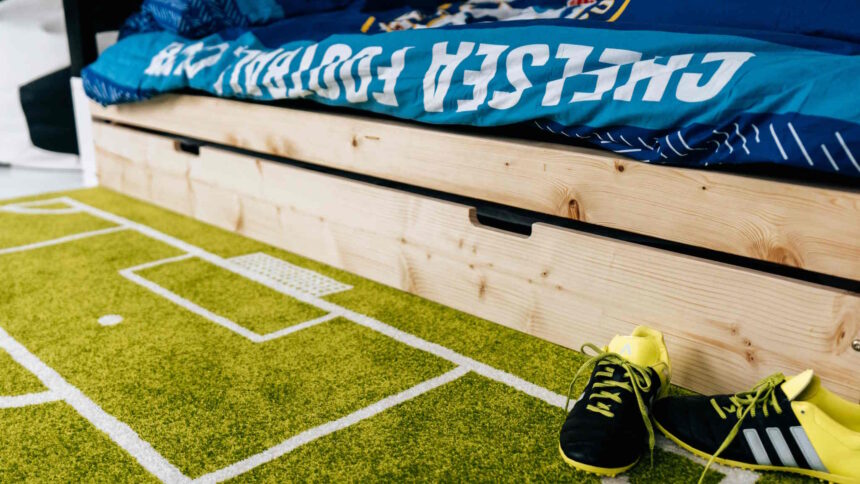 Drewniane łóżko bramka z wysuwaną szufladą na tle dywanu boiska i butów piłkarskich